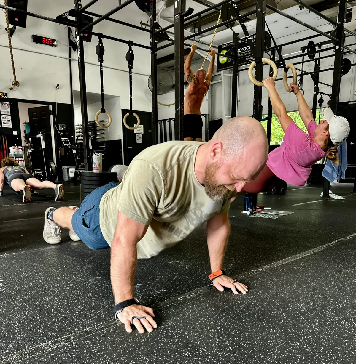 CrossFit Coordinate athlete mid-push-up on the gym floor during Murph, with teammates on the gymnastic rings in the background